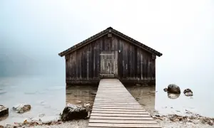 Drewniana szopa nad jeziorem, z pomostem prowadzącym do drzwi. Kompozycja otwarta, z mgłą w tle.