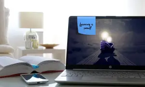 Laptop z ekranem pokazującym dwie postacie z żarówkami zamiast głów, siedzące na drewnianej kładce. Na ekranie notatka: "Learning is smart!". Obok otwarte książki i telefon.