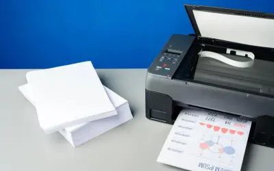 Stos białego papieru obok drukarki atramentowej, która właśnie drukuje dokument. Jaki papier do drukarki atramentowej wybrać, by uzyskać najlepsze rezultaty?