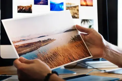 Dłonie trzymają wydruk krajobrazu. Różne rodzaje papieru do druku zdjęć są widoczne na ekranie komputera w tle.