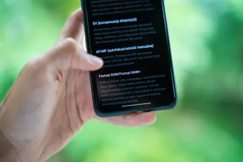 Dłoń trzyma smartfon, na ekranie którego widać opis formatu RAW. RAW to format pliku, który daje większą swobodę podczas obróbki zdjęć.