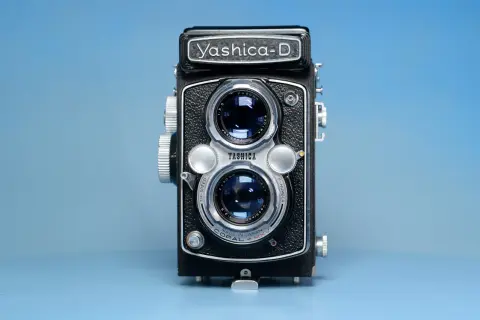 Czarno-srebrny aparat dwuobiektywowy Yashica-D na niebieskim tle.