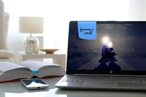 Laptop z ekranem pokazującym dwie postacie z żarówkami zamiast głów, siedzące na drewnianej kładce. Na ekranie notatka: "Learning is smart!". Obok otwarte książki i telefon.