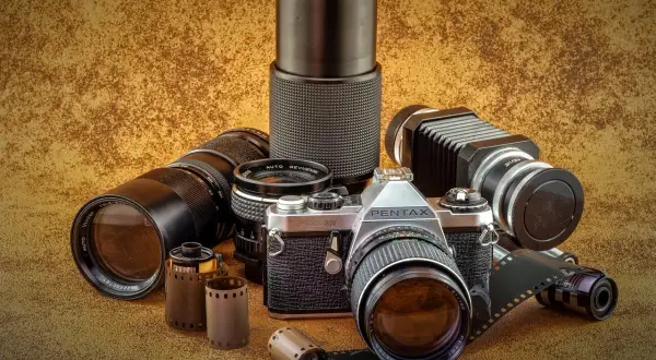Kolekcja różnych rodzajów aparatów fotograficznych i akcesoriów: lustrzanka Pentax, obiektywy, mieszek, filmy.