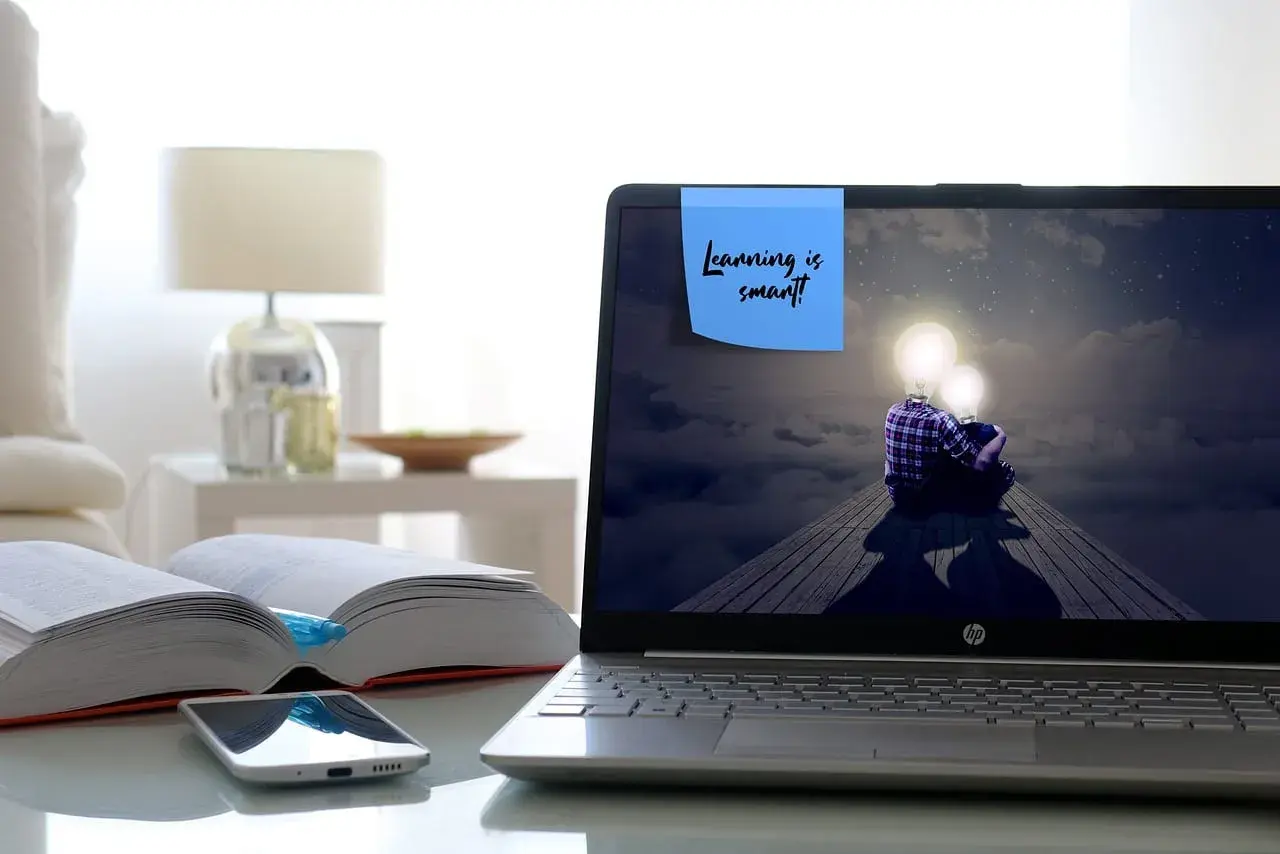 Laptop z ekranem pokazującym dwie postacie z żarówkami zamiast głów, siedzące na drewnianej kładce. Na ekranie notatka: "Learning is smart!". Obok otwarte książki i telefon.