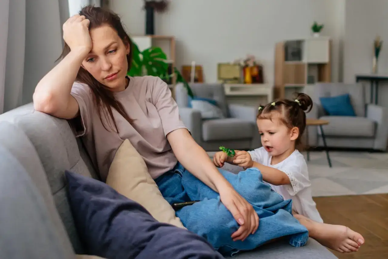 Zmęczona mama siedzi na kanapie, podczas gdy jej mała córeczka bawi się obok. Czasem z rodziną najlepiej wychodzi się na zdjęciu, ale nie zawsze.